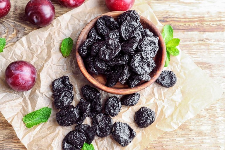 bowl of dried prunes surrounded with more prunes and leaf and plums on the paper at the table