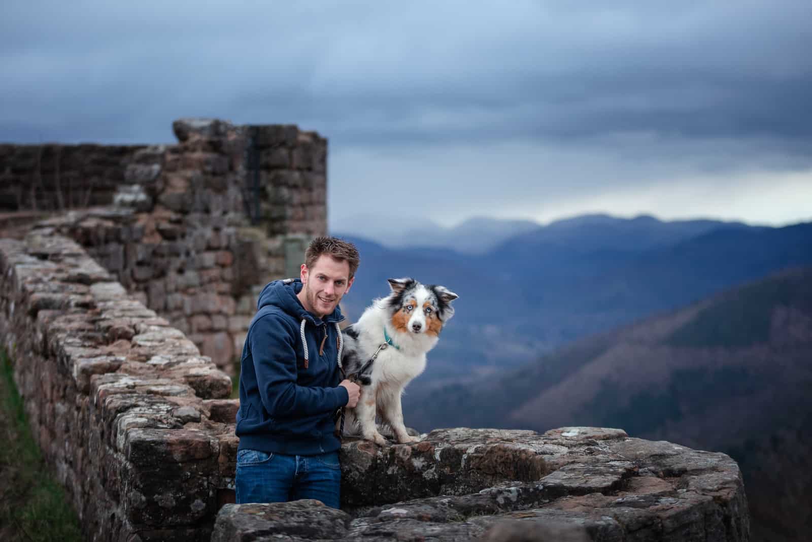 a man and his Australian Shepherd are sitting on a wall on a hill