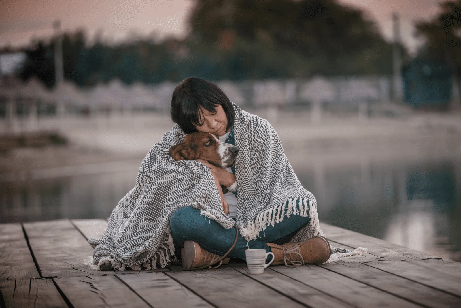 a woman sits on a pier and cuddles with a basset hound