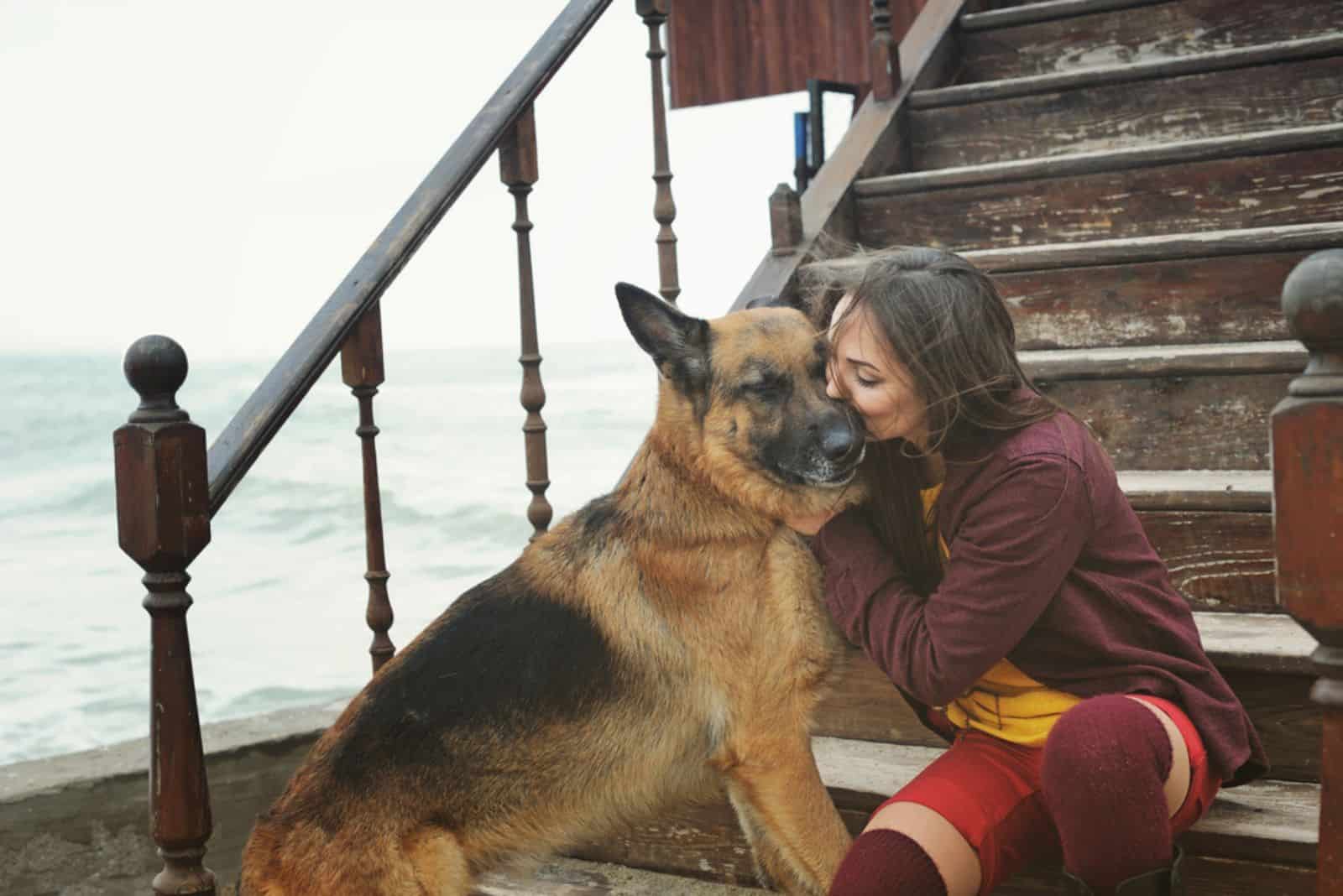Happy woman kissing her German shepherd dog on a stairs against sea
