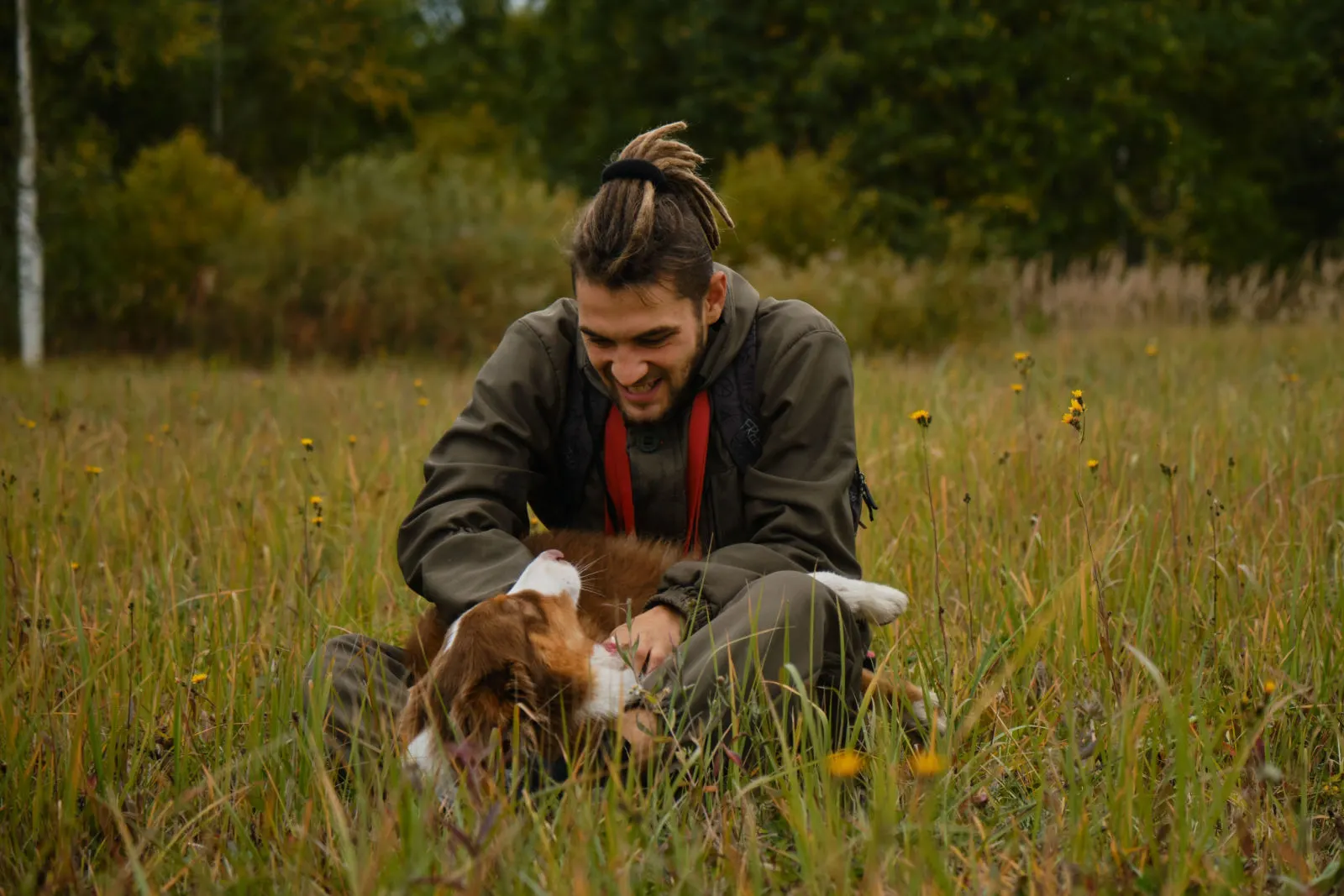 Young Caucasian man with dreadlocks and beard plays with Australian shepherd in autumn field.