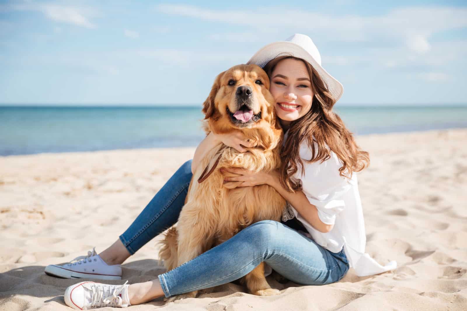 Cheerful pretty young woman in hat sitting and hugging her dog on the beach