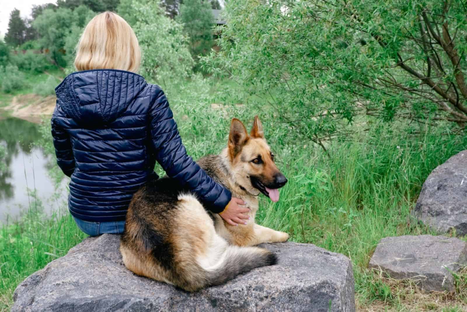 a woman with a german shepherd sits on a stone by the lake