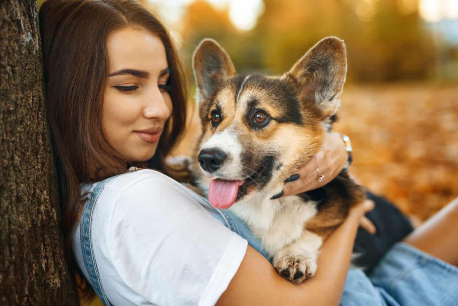 woman hugging corgi dog while sitting by tree