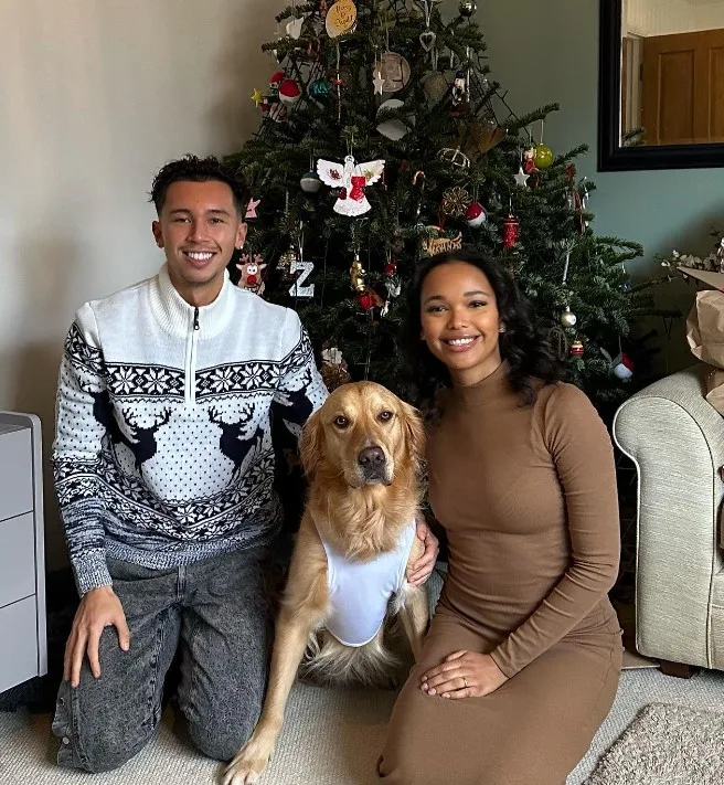a smiling loving couple with a golden retriever takes a picture next to the Christmas tree
