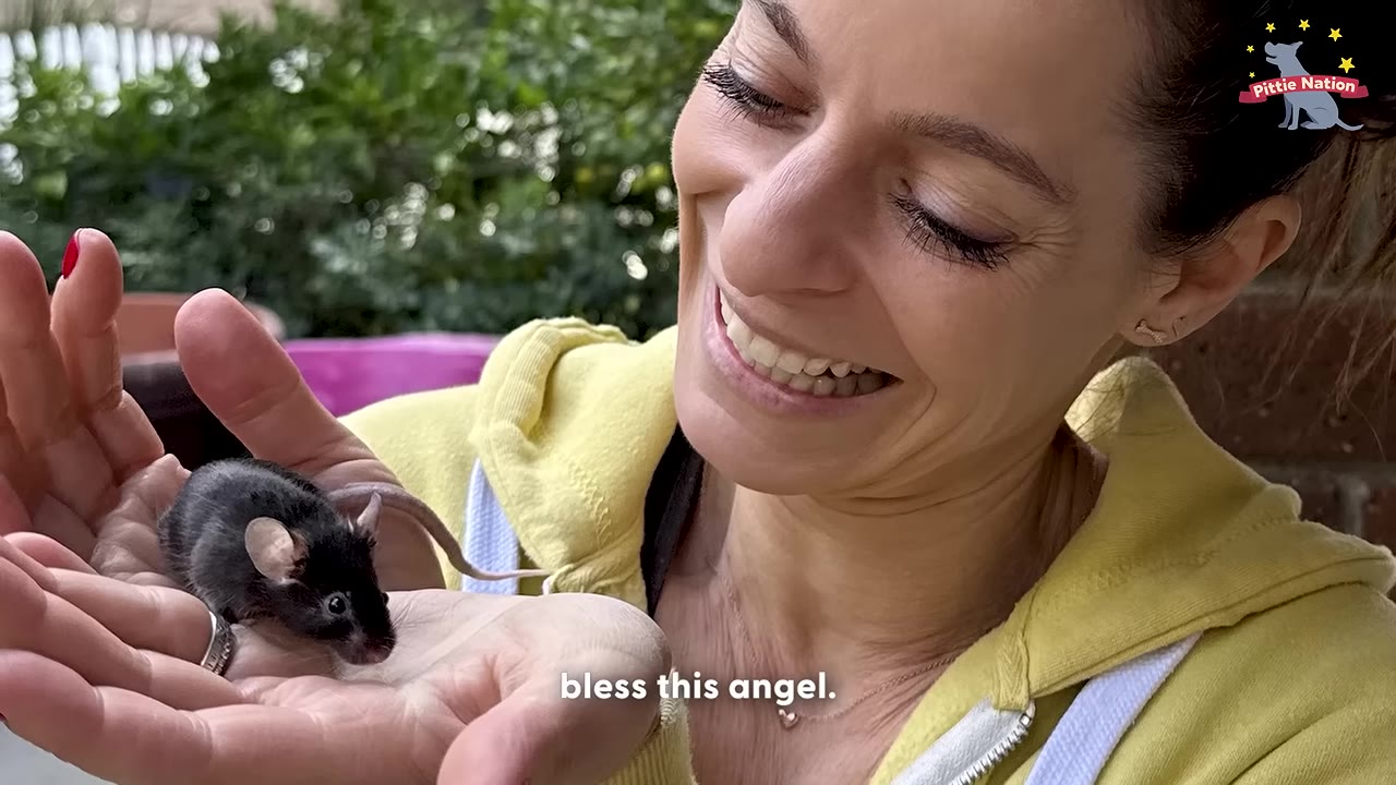 A close-up of a smiling woman holding a tiny black mouse in her cupped hands.