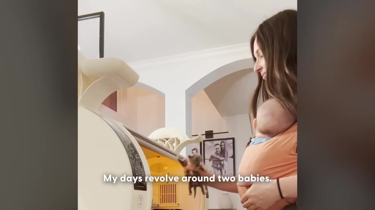 A woman wearing a baby in an orange wrap carrier holds a tiny puppy in her hand next to an incubator.