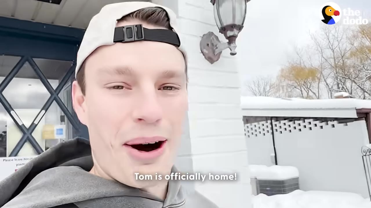 A man wearing a grey hoodie and a white baseball cap backwards smiles in front of a snowy background with the text 'Tom is officially home!'.