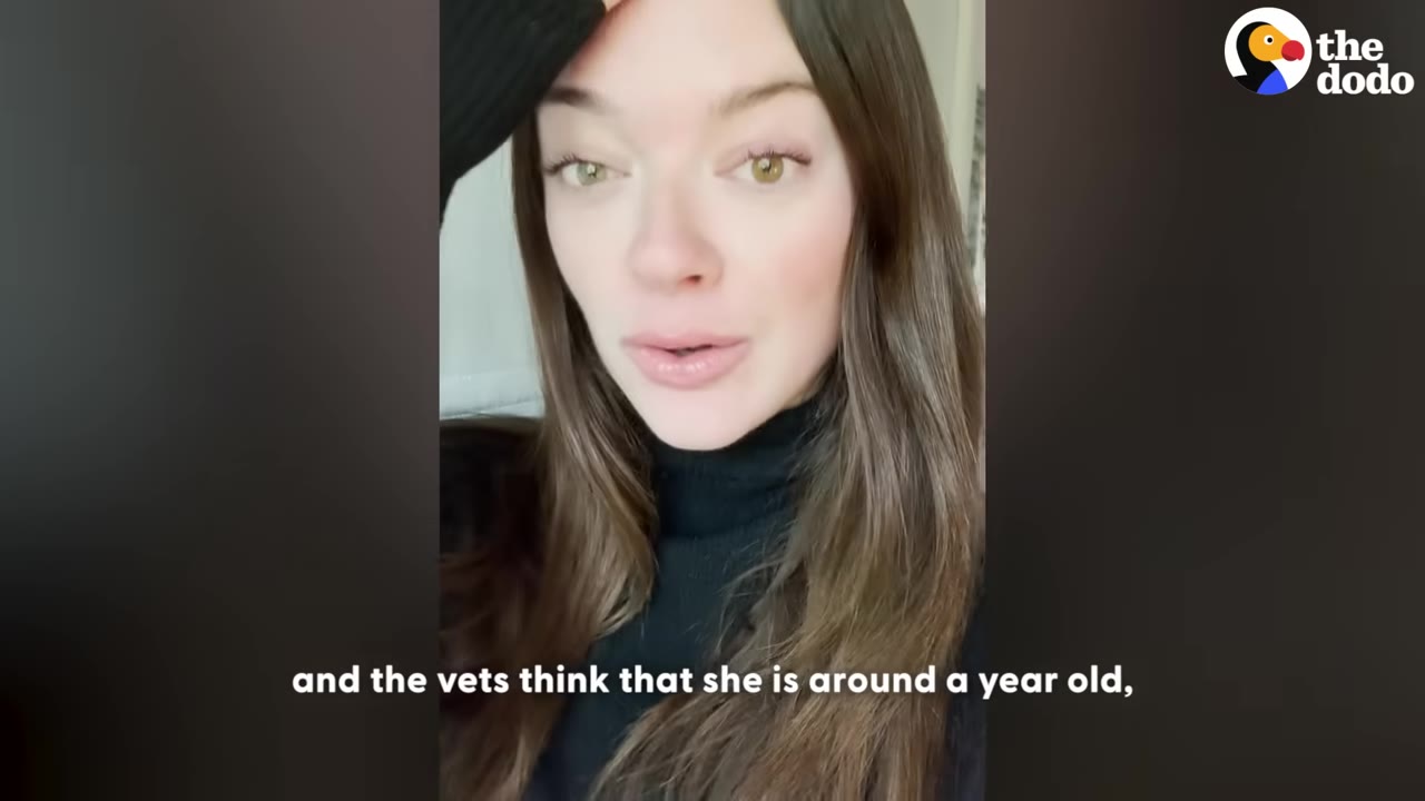 A close-up of a woman with long brown hair wearing a black turtleneck, looking directly at the camera with her hand raised to her forehead.