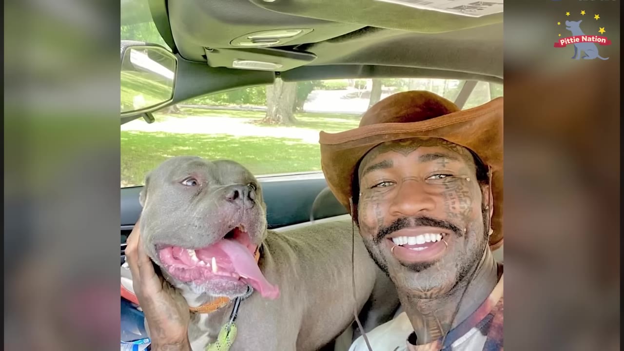 A smiling man wearing a brown cowboy hat sits in the driver's seat of a car next to a grey pit bull with its tongue out.