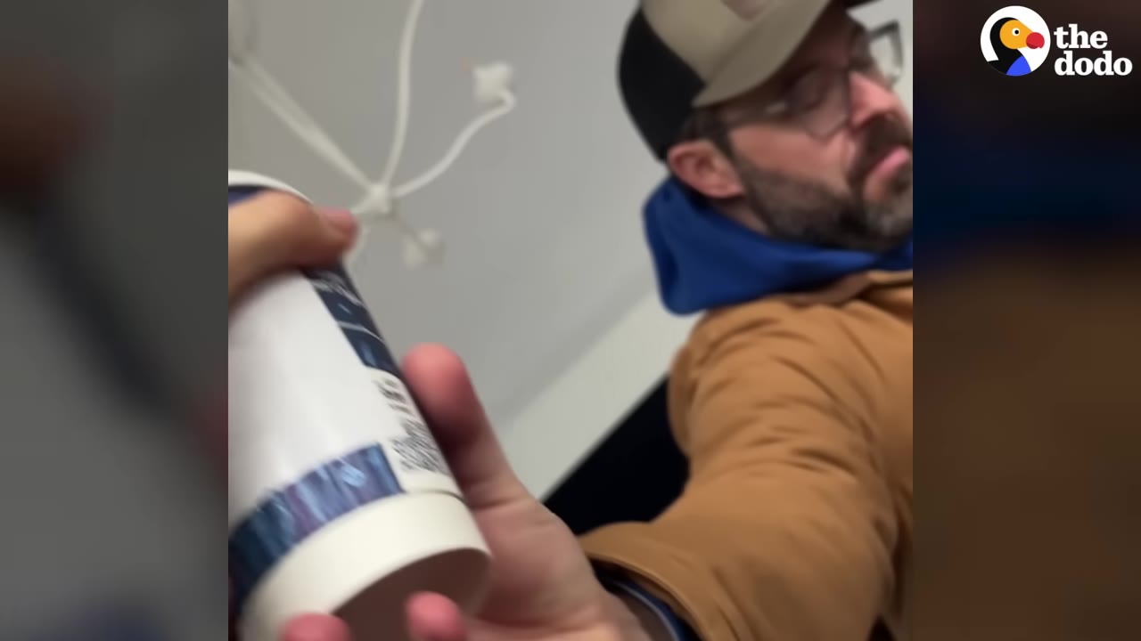 A man wearing a baseball cap and glasses looks tired while holding out a paper coffee cup.