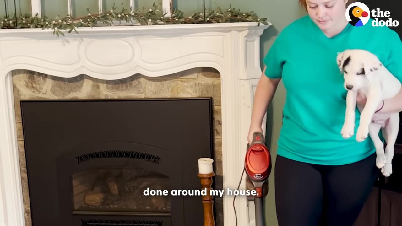 A woman in a green t-shirt holds a white puppy with black spots in one arm while holding a red vacuum cleaner in her other hand.
