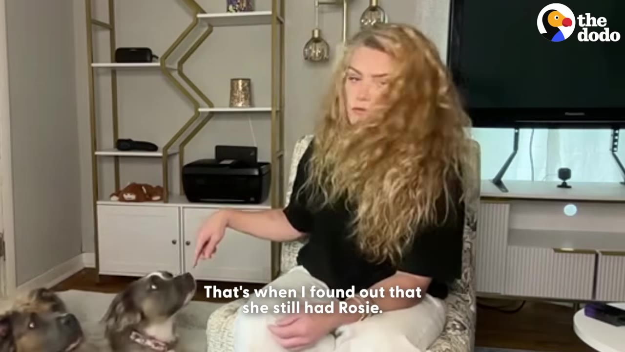 A woman with long blonde curly hair sits in a chair, pointing towards a dog looking up at her, with on-screen text reading 'That's when I found out that she still had Rosie.'