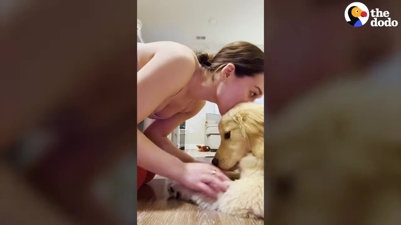 A woman leans down to kiss the head of a golden retriever puppy sitting on the floor inside a house.