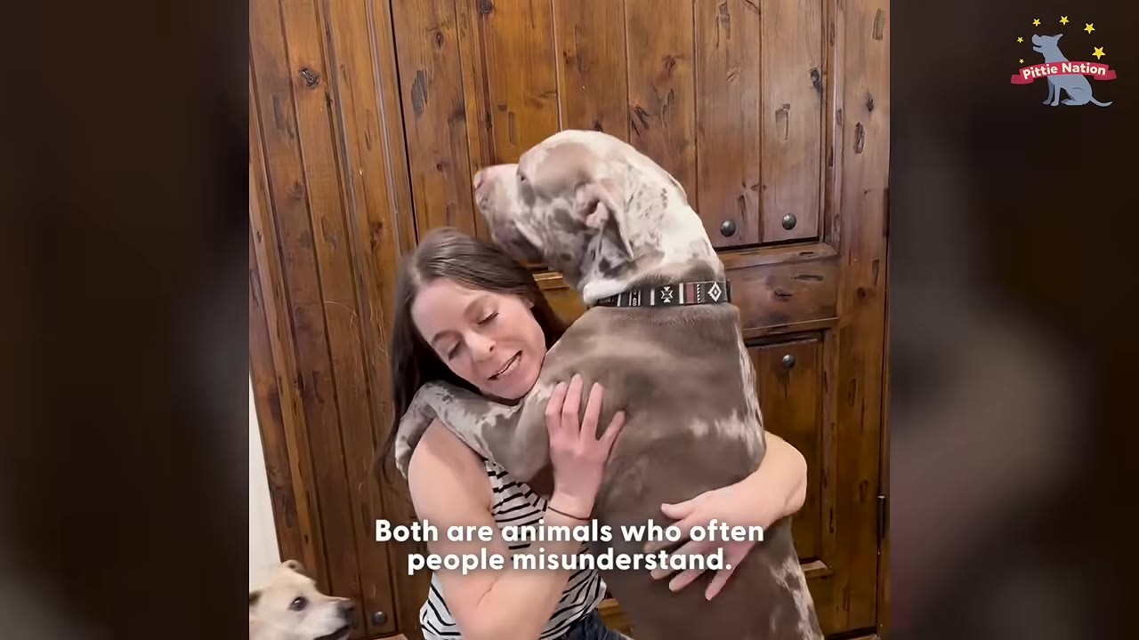 A woman with long brown hair smiles as she hugs a large, spotted pit bull dog standing on its hind legs against a wooden door.