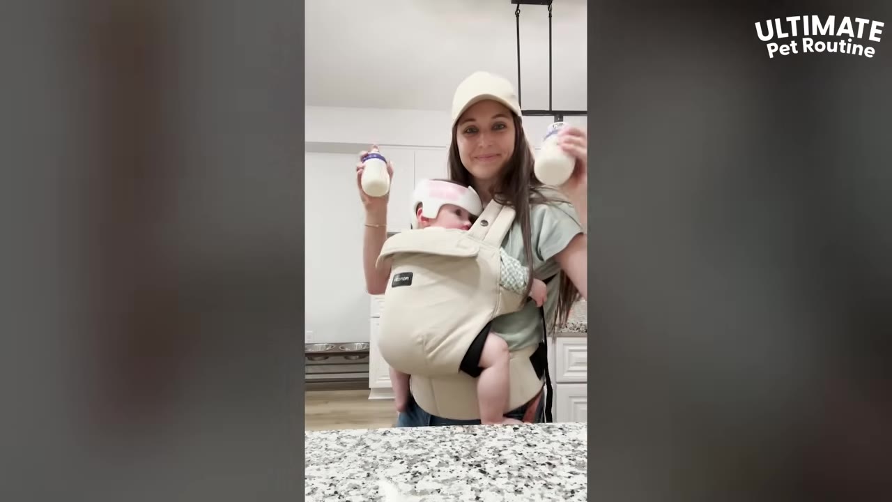 A smiling woman wearing a baseball cap holds a baby in a front carrier while holding up two baby bottles in a kitchen.