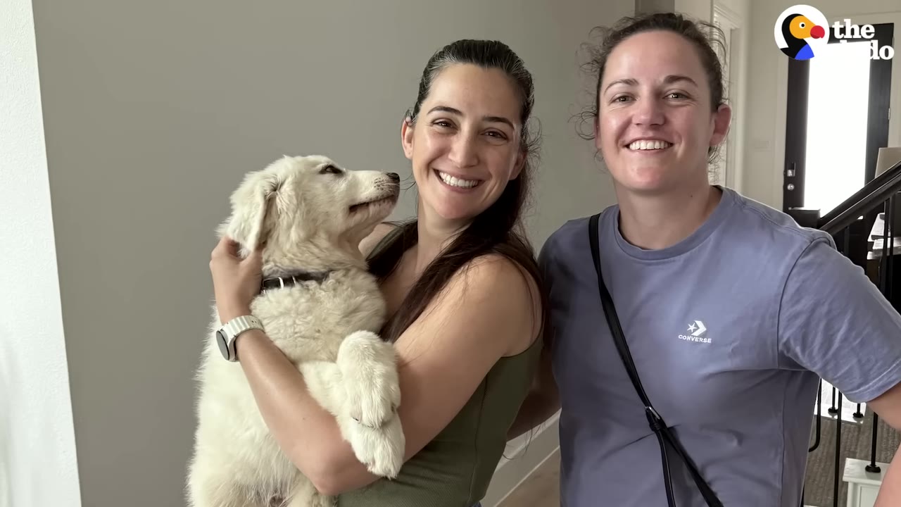 Two smiling women stand indoors, with one holding a fluffy white puppy that looks up at her.