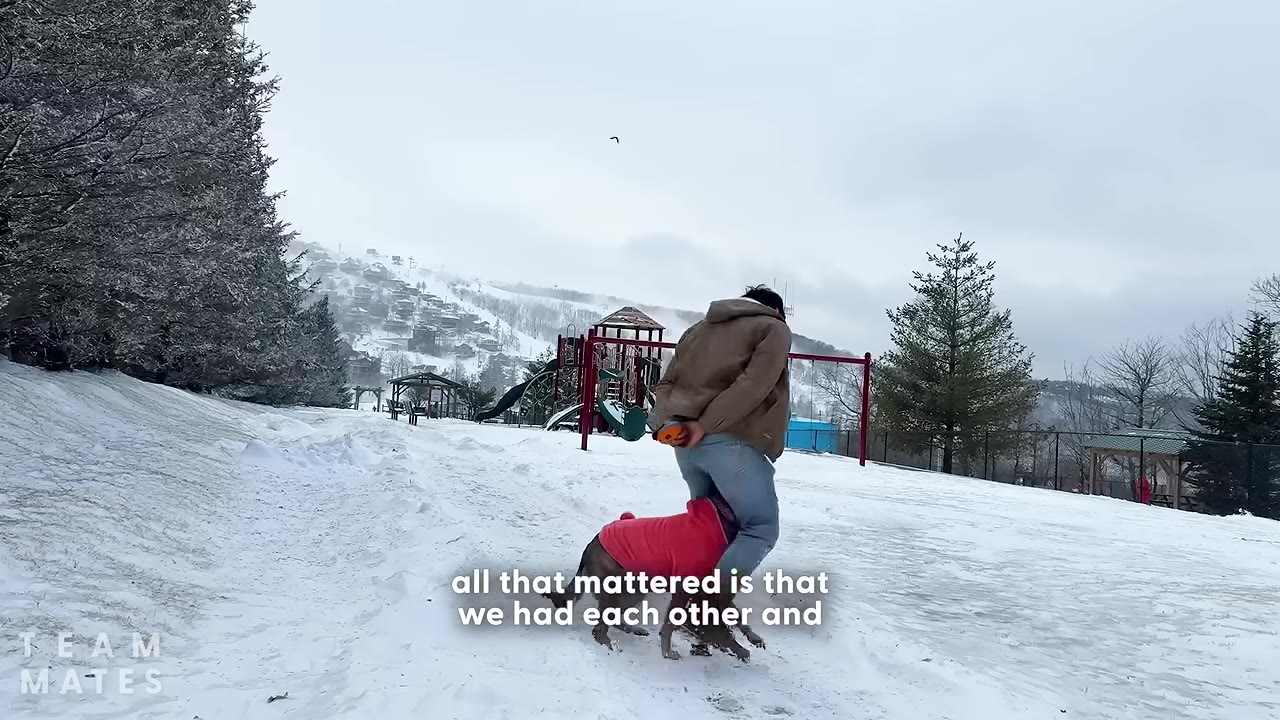 A dog wearing a pink sweater runs playfully around a man's legs in a wide, snow-covered park with a playground in the background.