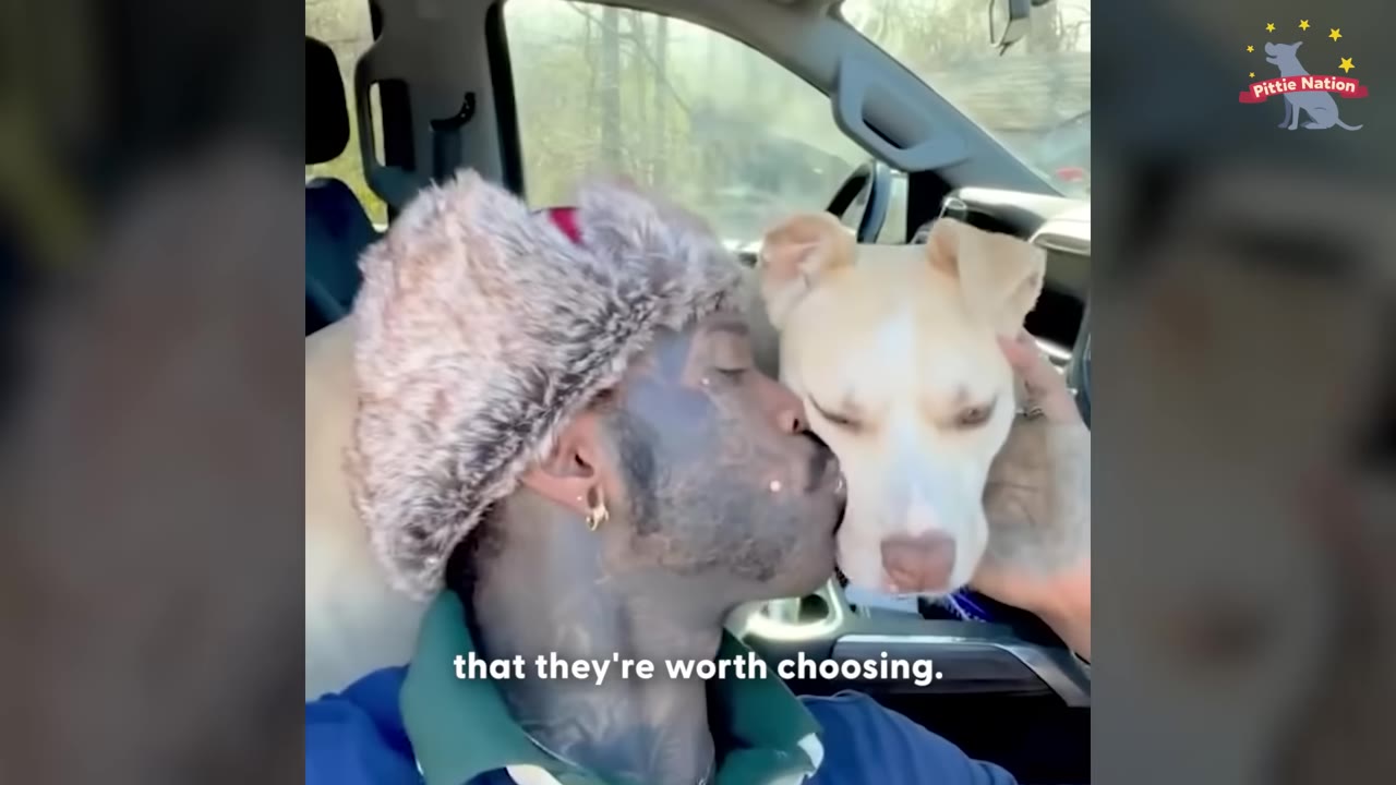 A person wearing a furry winter hat kisses a light-colored dog on the cheek inside a vehicle.