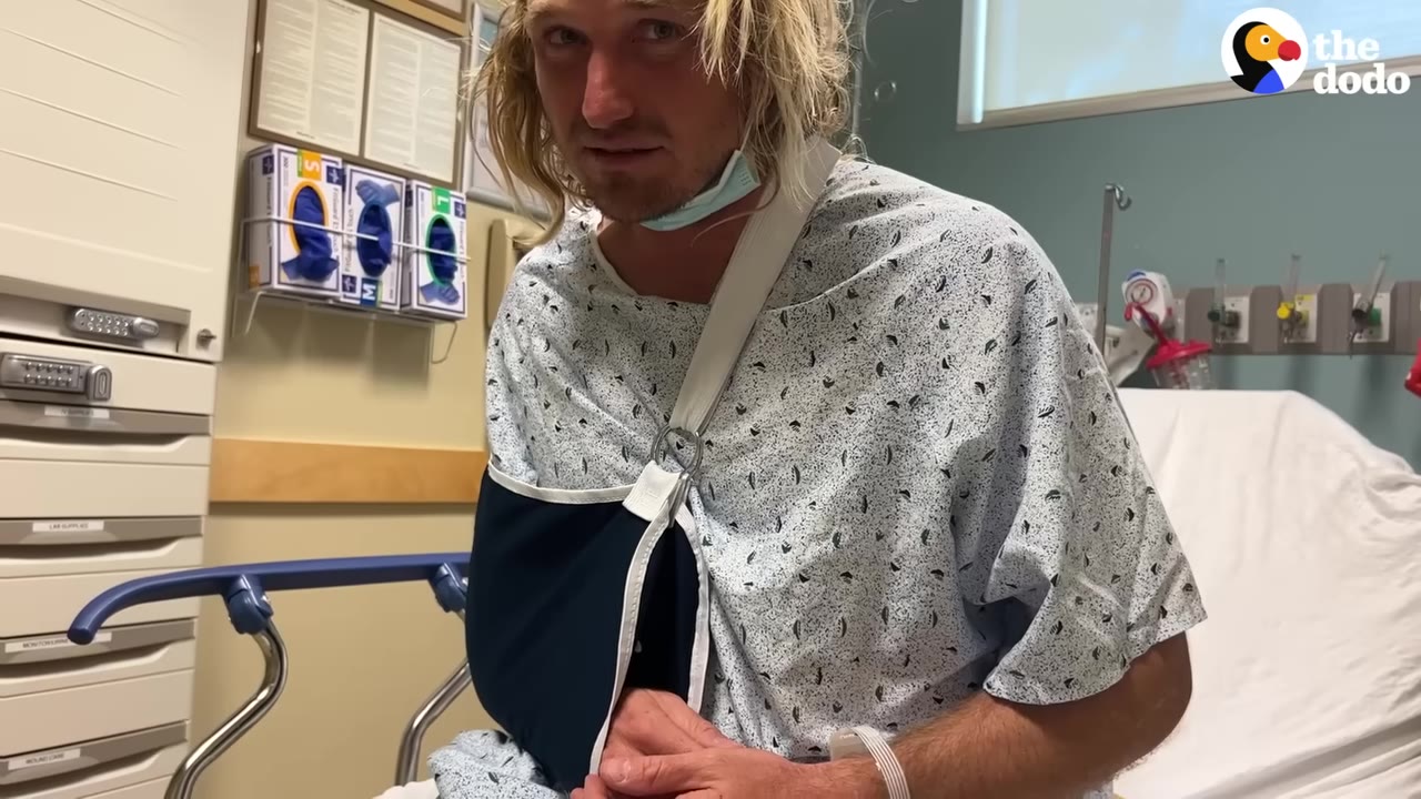 A man with blonde hair wearing a hospital gown and a black arm sling sits on a hospital bed.