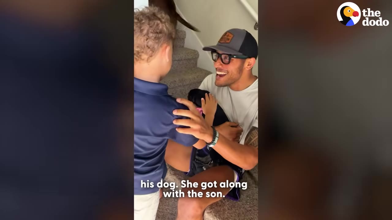 A smiling man wearing a baseball cap and glasses sits on carpeted stairs holding a small black puppy, while a young boy stands beside him.