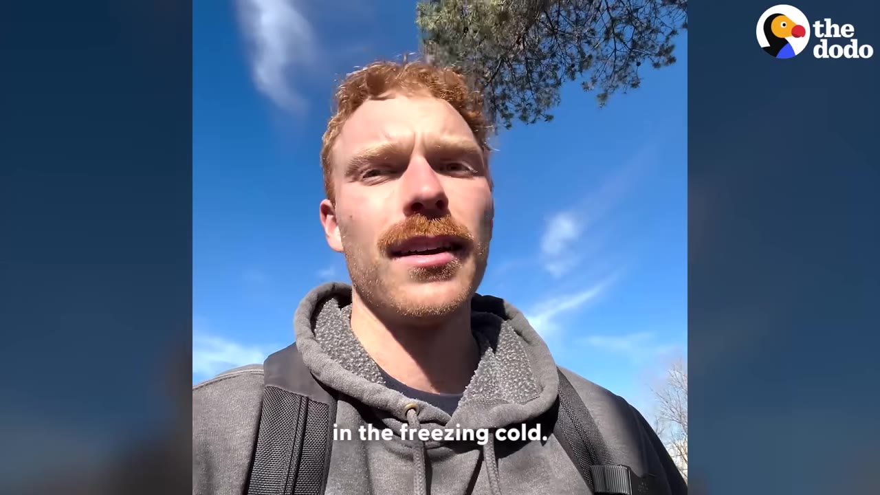 A man with red hair and a mustache wearing a grey hoodie speaks to the camera against a blue sky background with the caption 'in the freezing cold.'