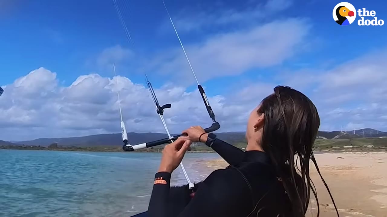 A woman in a black wetsuit stands on a sandy beach holding a kitesurfing control bar, looking up at the blue sky with white clouds.