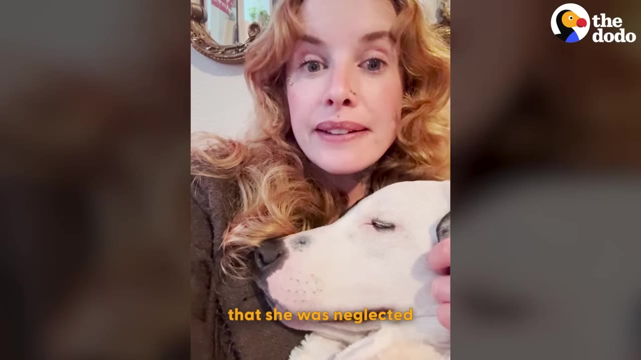A woman with wavy blonde hair looks directly at the camera while gently holding a white dog with black spots that is resting its head on her chest.