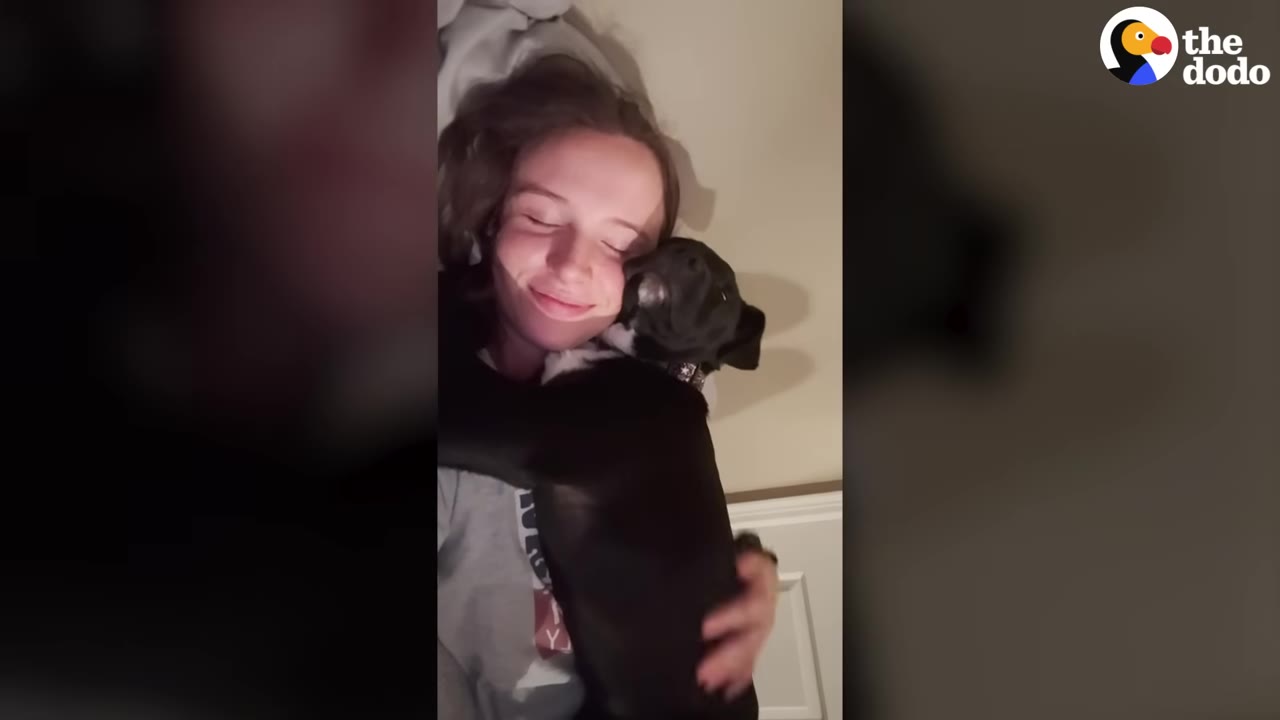 A smiling woman lying down and hugging a black dog close to her chest.