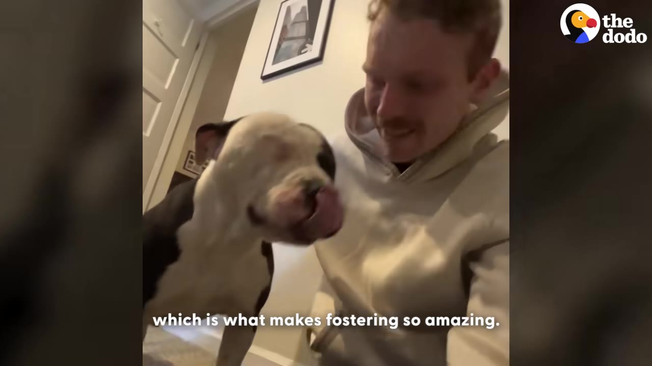 A smiling man in a beige hoodie sits next to a black and white dog who is licking its nose.
