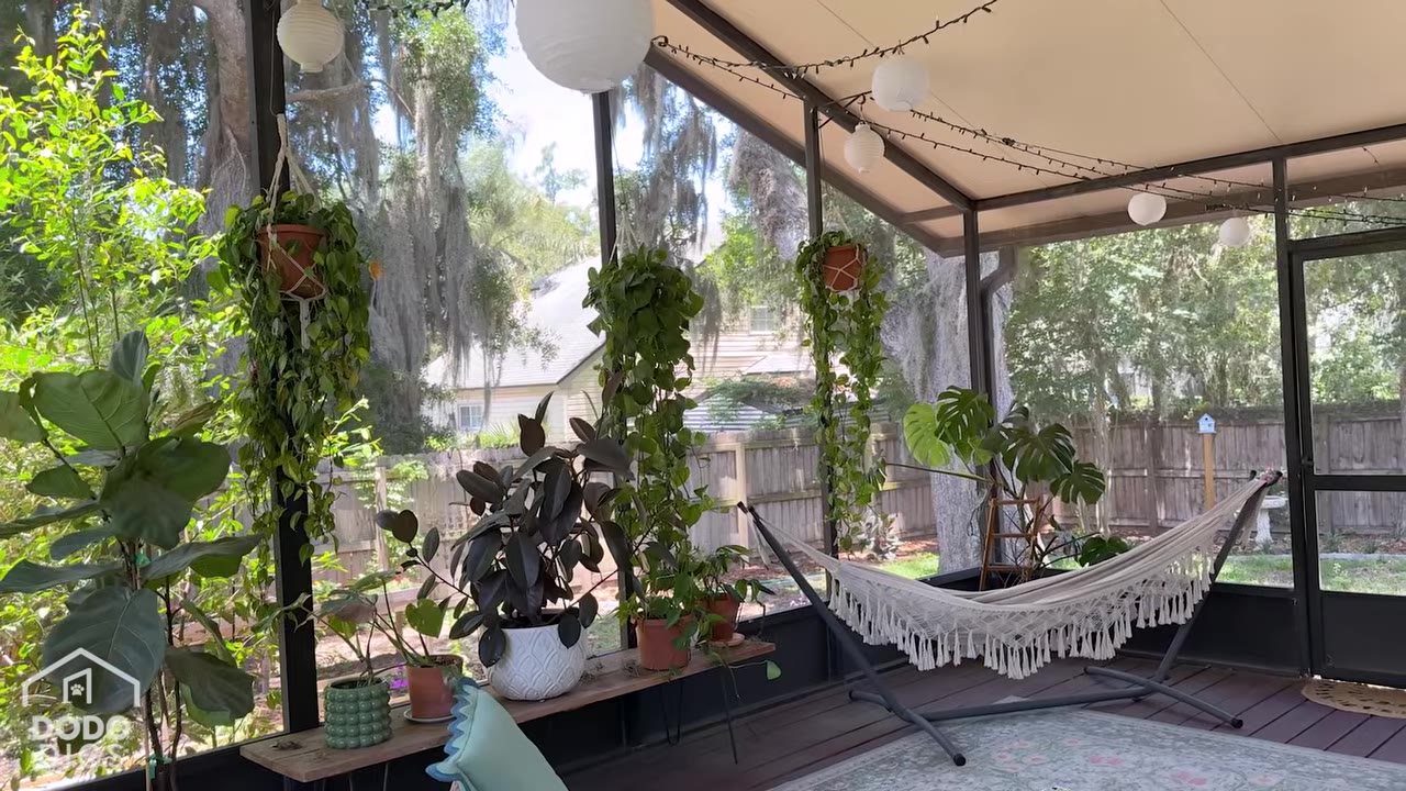 A peaceful screened-in porch filled with lush green hanging and potted plants, featuring a white woven hammock under string lights.