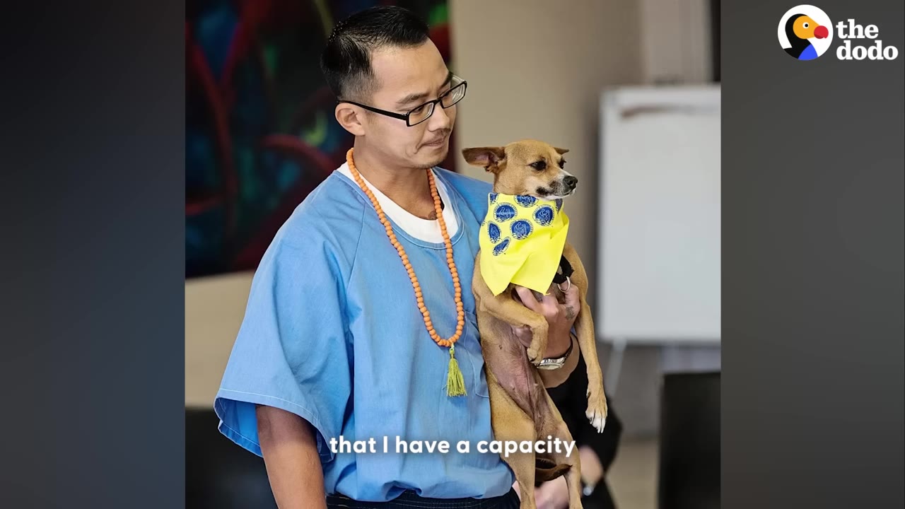 A man wearing glasses, a blue shirt, and an orange beaded necklace holds a small brown dog wearing a yellow bandana.