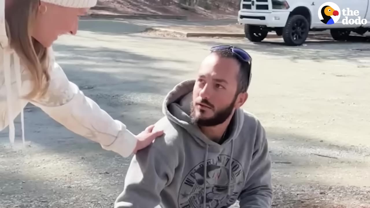 A woman in a white beanie places her hand on the shoulder of a bearded man in a grey hoodie, who looks up at her with a surprised expression.