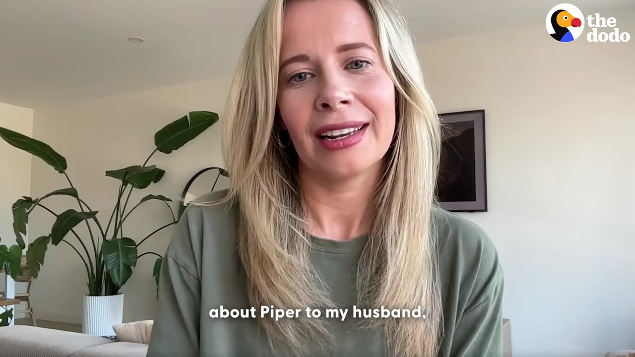 A blonde woman in a green shirt speaks directly to the camera in a brightly lit room.