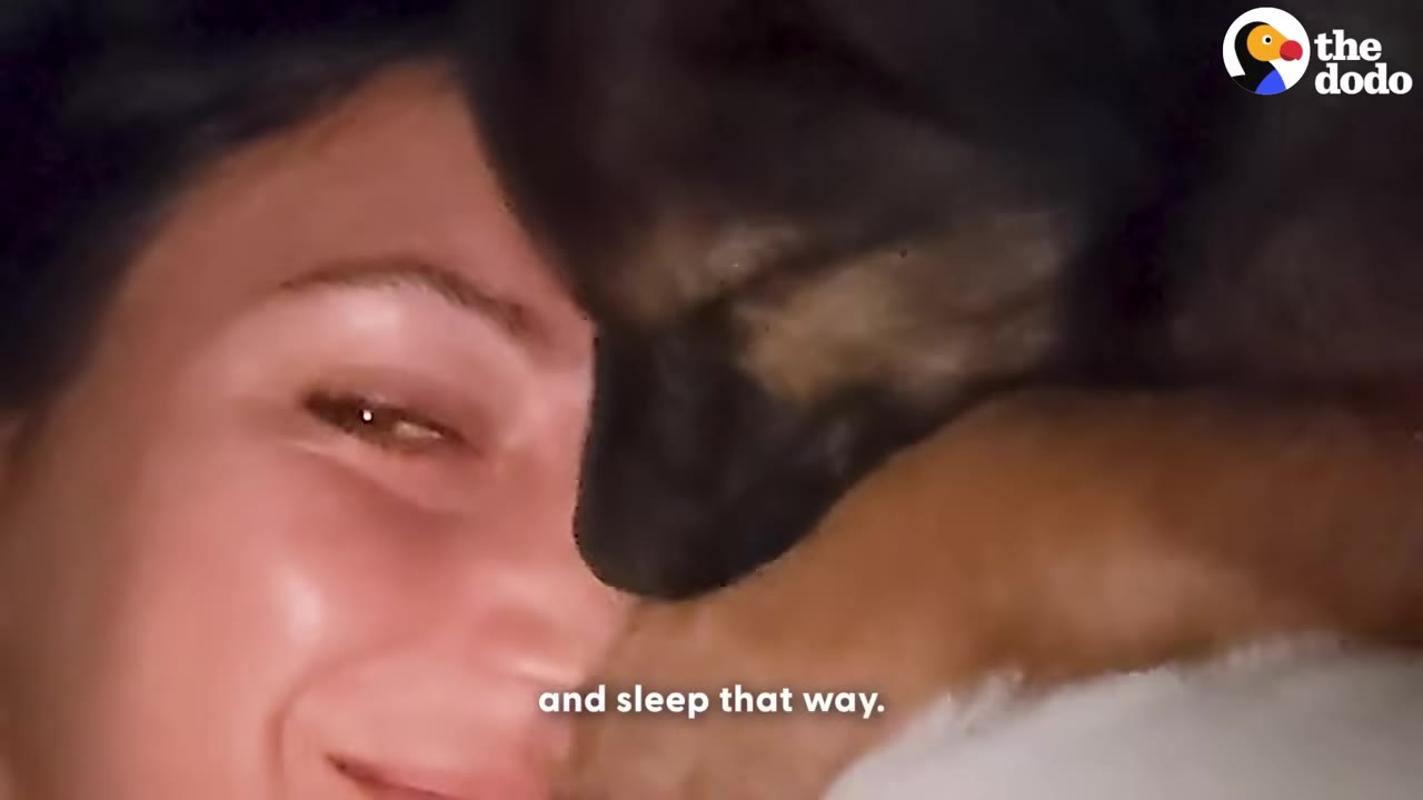 A close-up of a smiling woman cuddling with a small dark-furred puppy resting its head against her face.