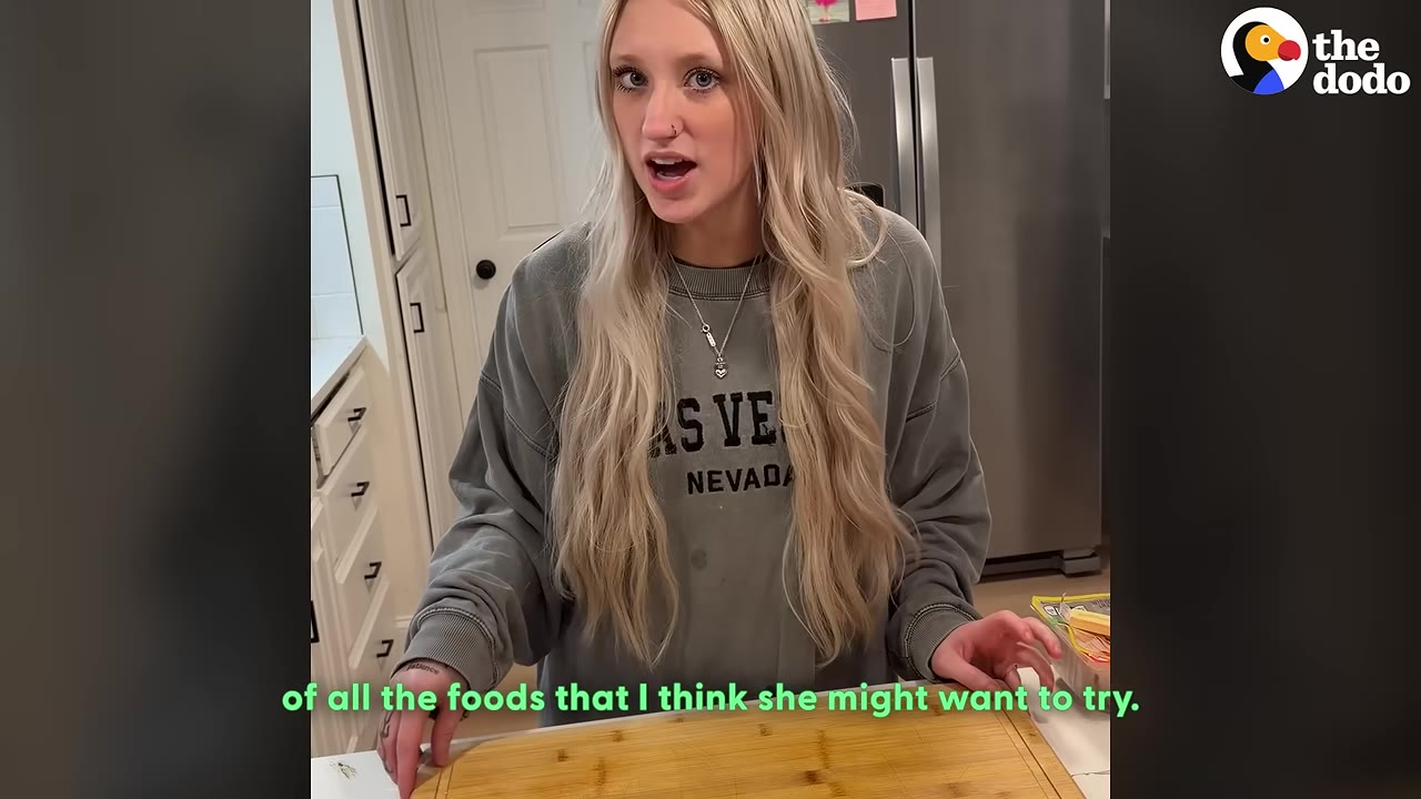 A young woman with long blonde hair stands in a kitchen behind a wooden cutting board, explaining her plan to prepare different foods.