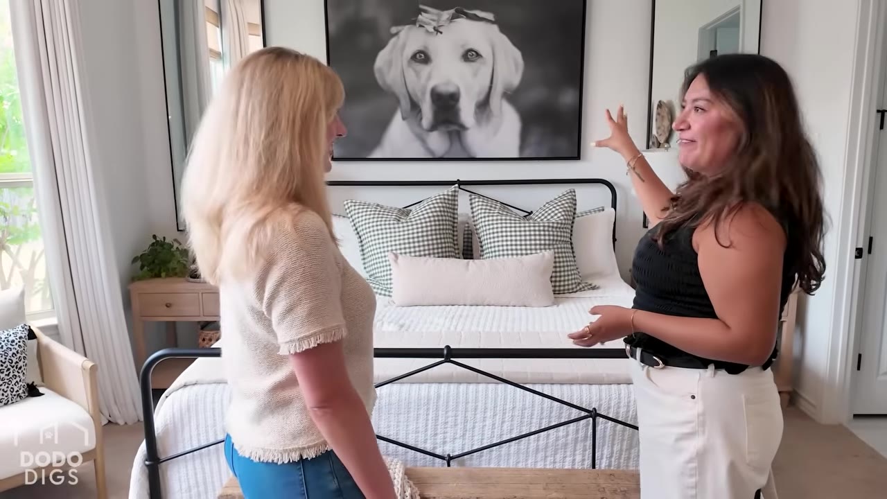 Two women stand in a bedroom looking at a large, black-and-white portrait of a yellow Labrador retriever wearing a flower crown, which hangs directly above a bed.