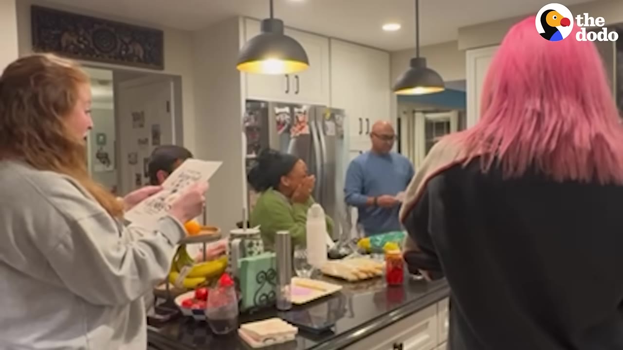 A woman in a green sweater covers her mouth in surprise in a kitchen while others hold up a flyer.