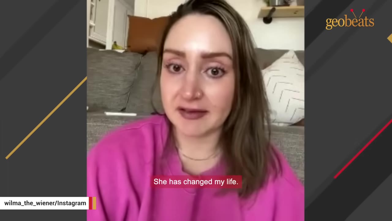 A close-up of a woman with brown hair wearing a pink sweatshirt, looking directly at the camera with a reflective expression, with a subtitle that reads 'She has changed my life.'