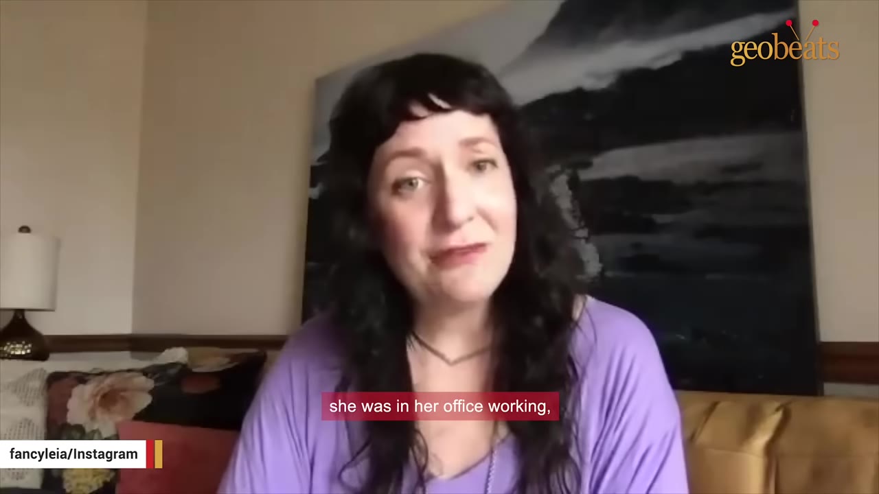 A woman with dark hair and a purple shirt speaking in front of a painting.
