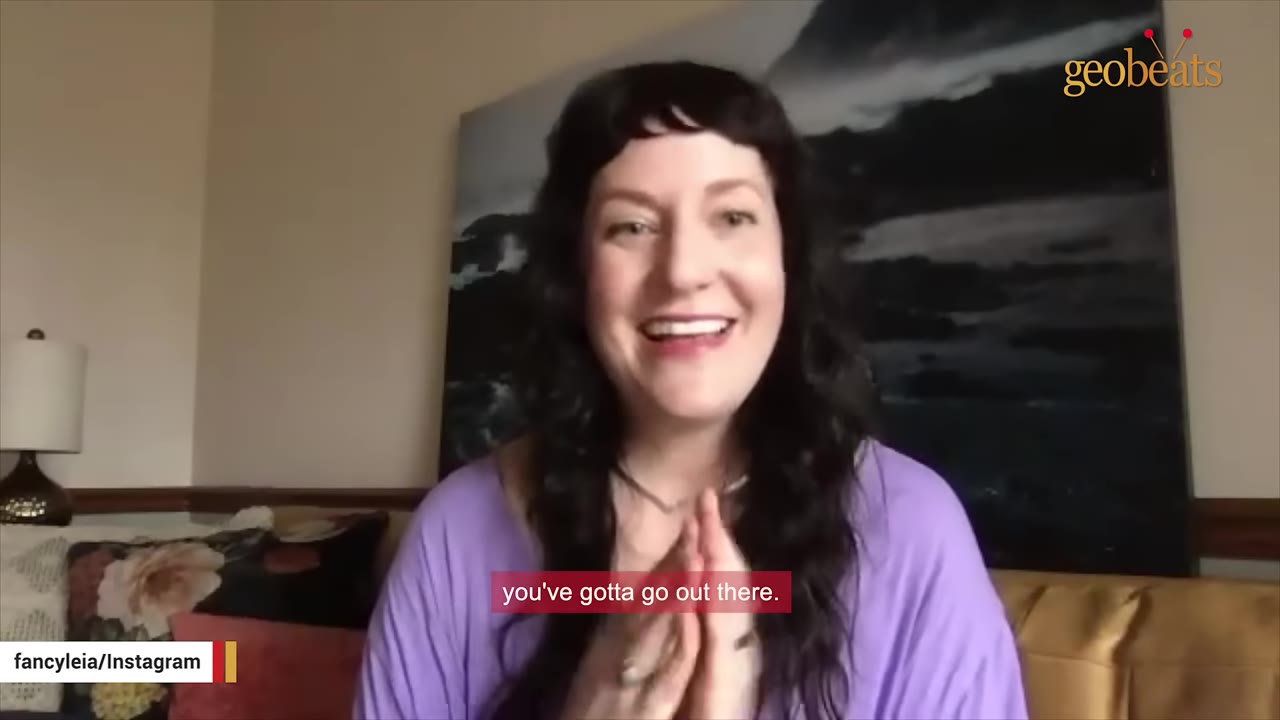 A woman with long dark hair and a purple shirt smiles while speaking during an interview.